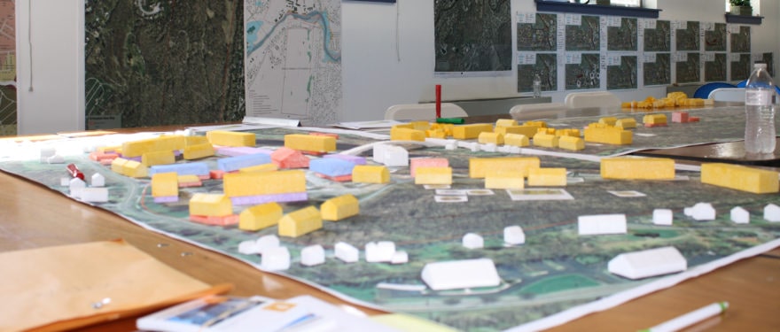 On a tabletop, small yellow and white model buildings are spread out on a green overhead map of a town as part of a public planning process workshop run by Dodson & Flinker.